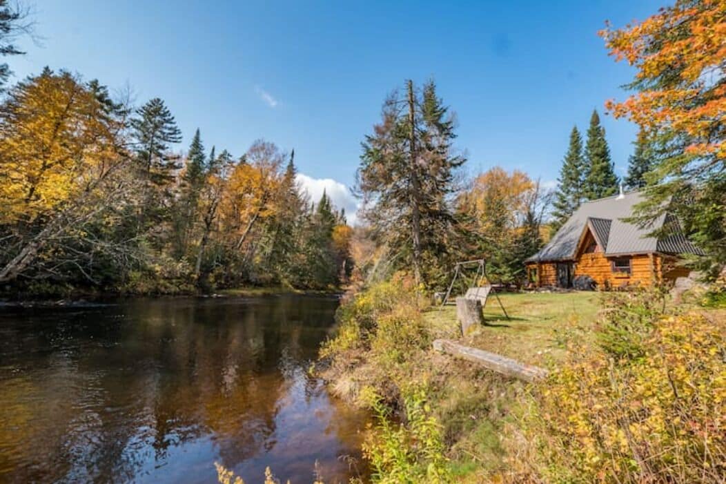 A cabin by a lake with autumn foliage, surrounded by trees and a clear sky.