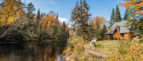 A cabin by a lake with autumn foliage, surrounded by trees and a clear sky.