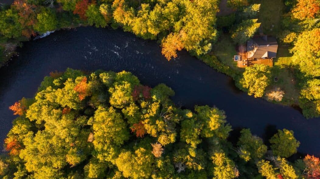 Vue aérienne d’une maison entourée d’arbres aux feuillages denses, typiques de l’automne, avec une rivière et un petit quai.