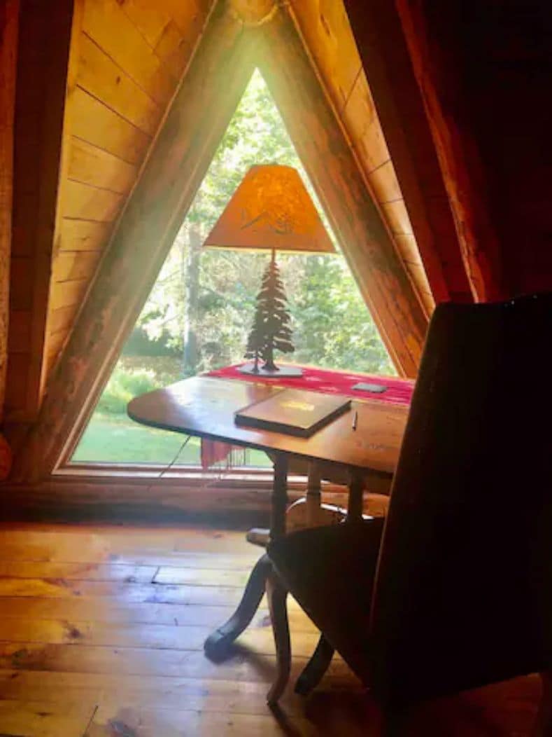 A room with a wooden floor, a table with a lamp, and a view of greenery outside.