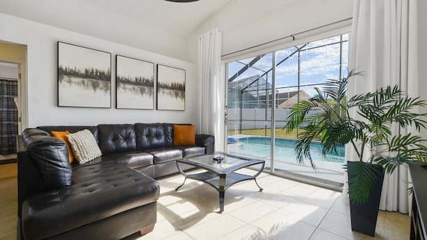A modern living room with a leather sofa, a glass coffee table, and a large window overlooking a swimming pool. - Private Heated Pool, Game Room, 8 mi to Disney, Walk to #GKTW! (Kissimmee)