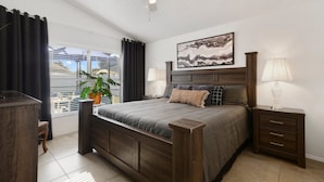 A bedroom with a large bed, a ceiling fan, a nightstand with a lamp, a window with curtains, and a plant. - Private Heated Pool, Game Room, 8 mi to Disney, Walk to #GKTW! (Kissimmee)