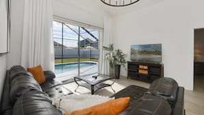 A modern living room with a large window, a pool view, a sofa, a TV, and a ceiling light fixture. - Private Heated Pool, Game Room, 8 mi to Disney, Walk to #GKTW! (Kissimmee)