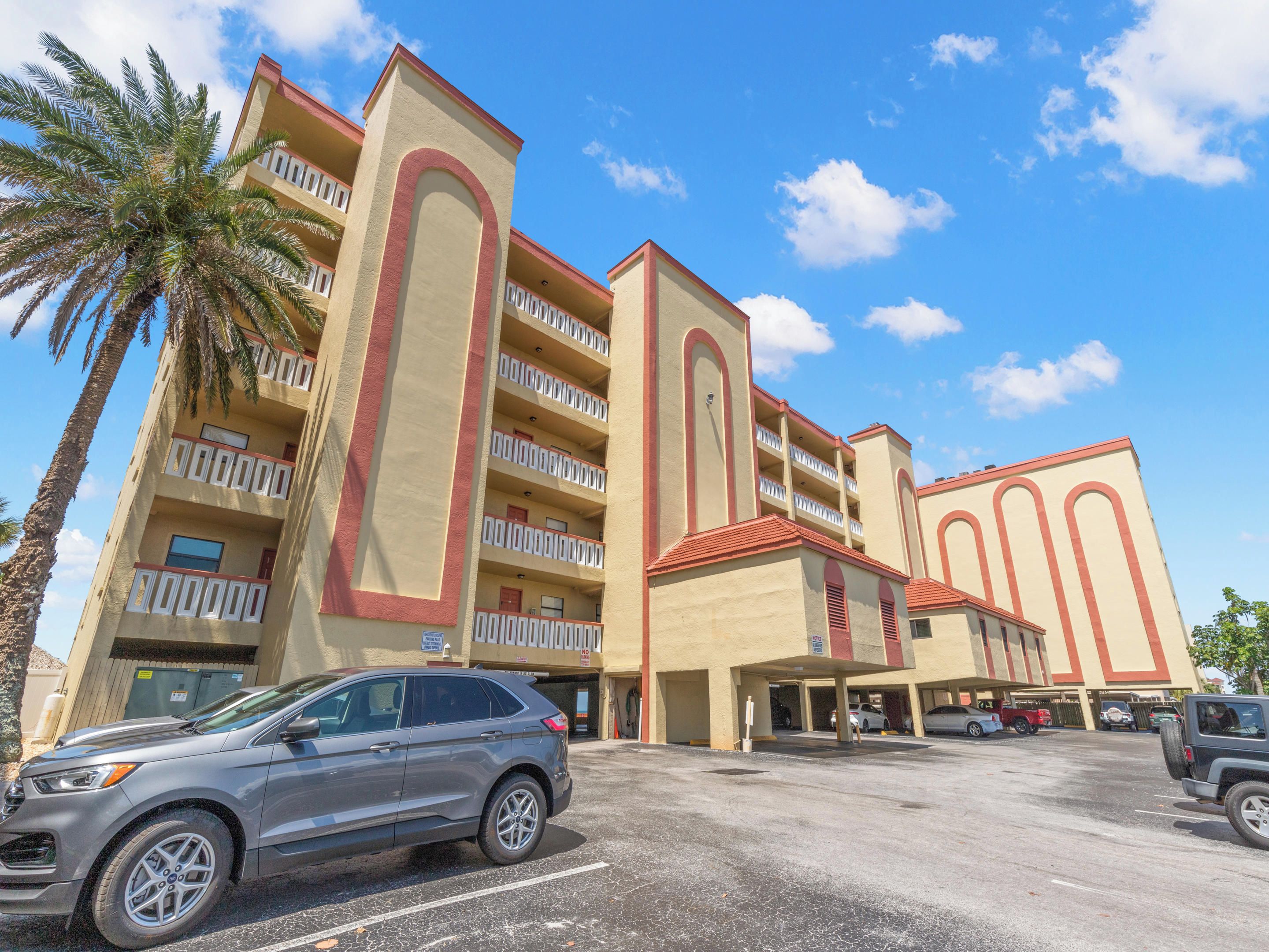 A multi-story hotel with a parking lot, featuring a modern architectural design with balconies and a clear blue sky.