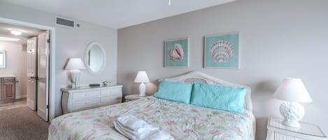 Une chambre à coucher comprenant un lit, deux tables de chevet, un ventilateur de plafond, un miroir et deux tableaux encadrés accrochés au mur.