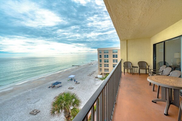 A balcony with a view of a beach and ocean, featuring outdoor seating and a building in the background.