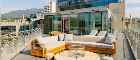 Una terrazza sul tetto con una moderna area salotto esterna, un edificio con pareti in vetro e vista sullo skyline cittadino e sulle montagne sullo sfondo.