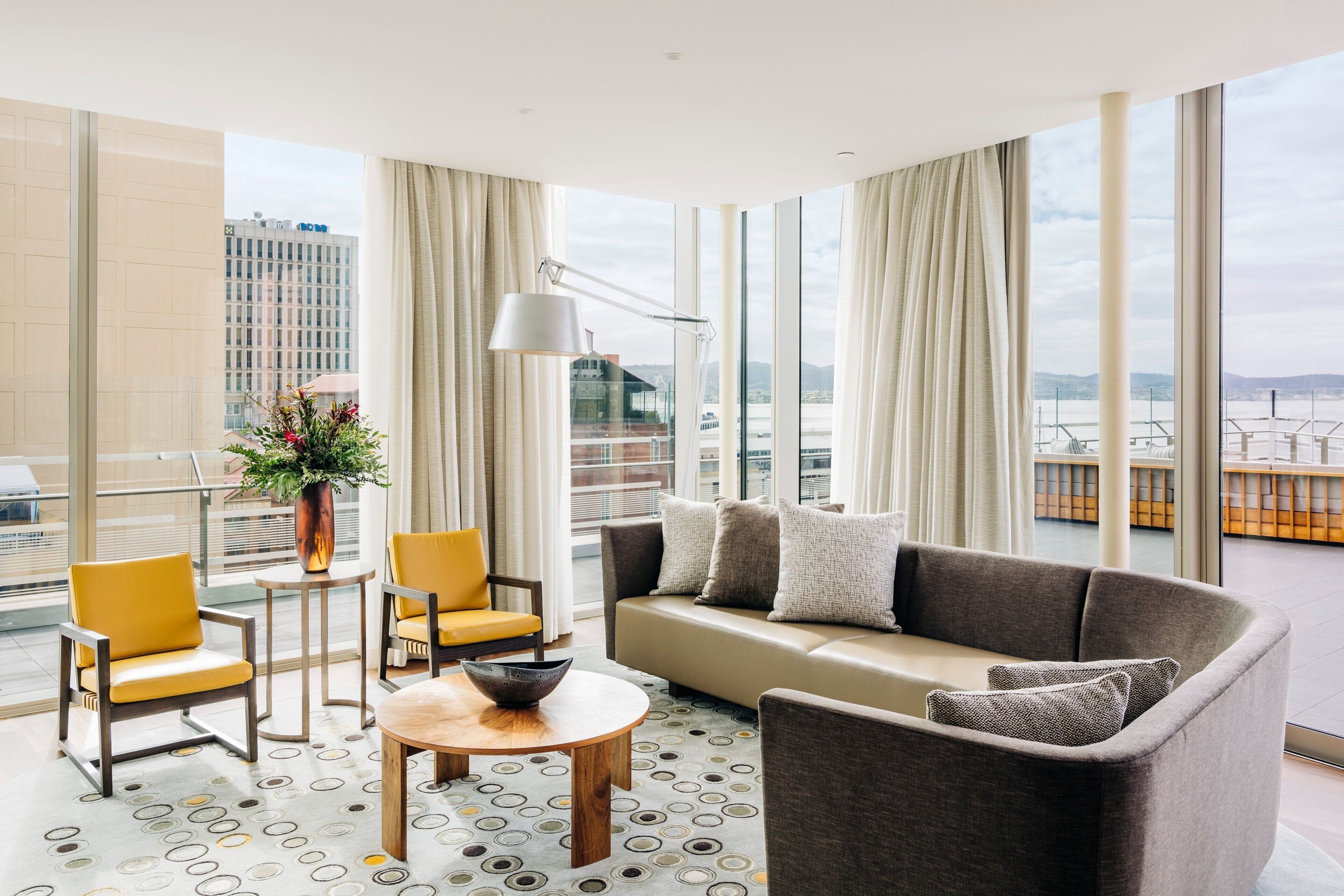 Une chambre d’hôtel moderne dotée d’une grande fenêtre, d’un canapé, de deux fauteuils, d’une petite table et d’un lampadaire.