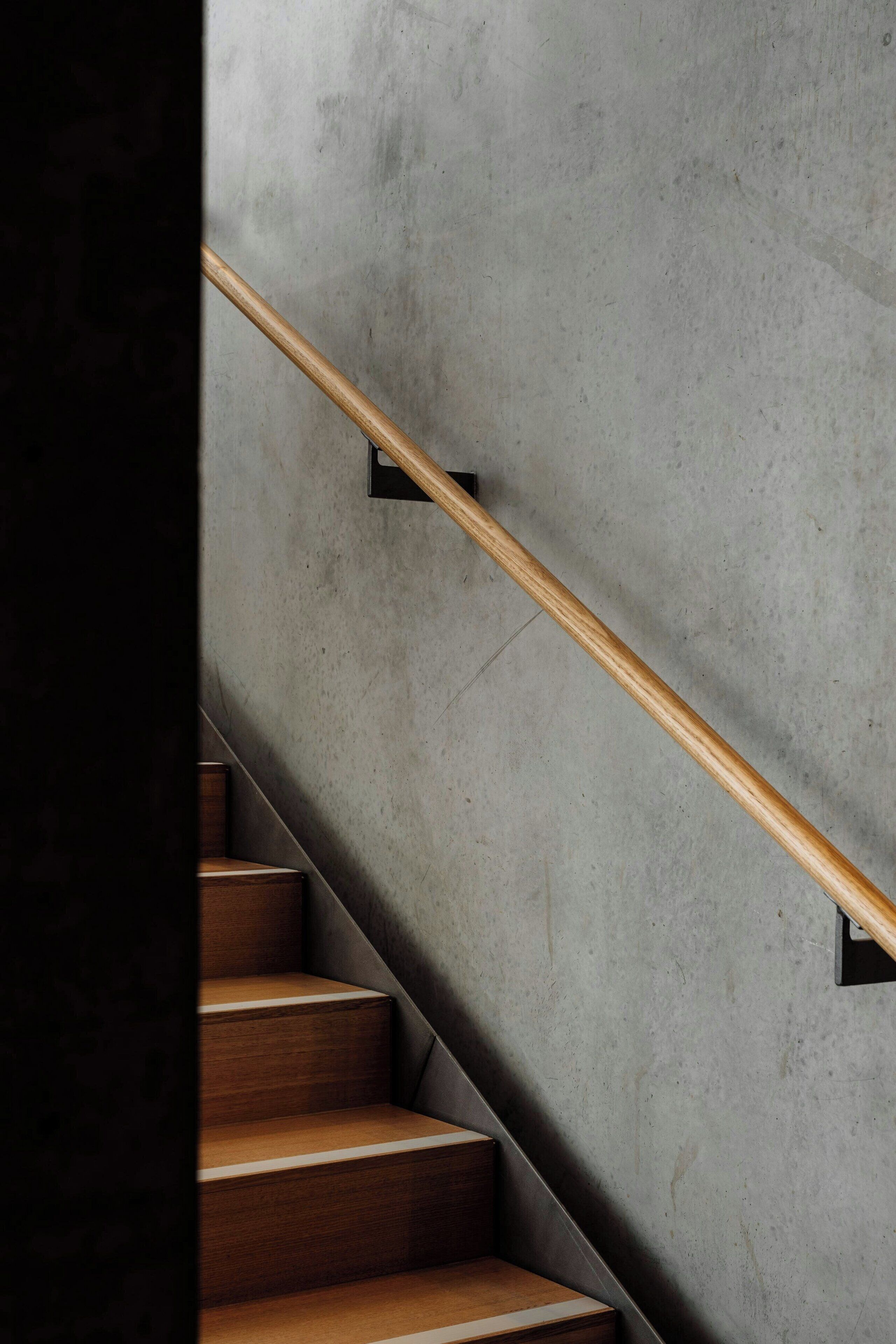 Una scala con gradini in legno e una ringhiera di legno, addossata a un muro in cemento.