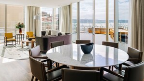 A modern hotel room with a large round table, comfortable seating, a sofa, and a view of the city through large windows.
