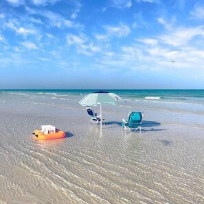 A beach with clear skies, a floating inflatable, two beach chairs, and a striped umbrella.