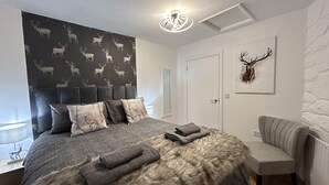 A modern bedroom with a large bed, a headboard with a deer pattern, a grey furry throw, and a grey armchair. - Solway Cottage (Annan)