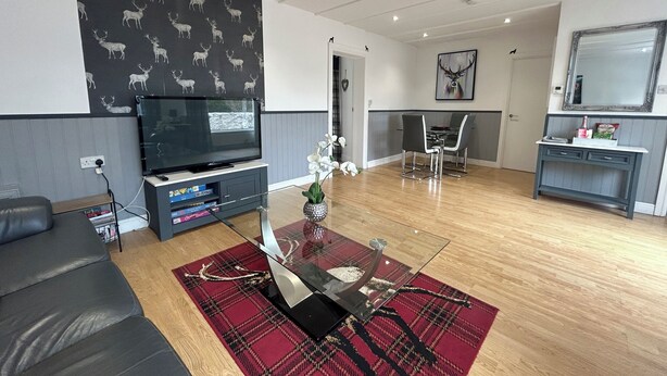 A modern living room with a flat-screen TV, a glass coffee table, a red plaid rug, a grey sofa, and a dining area with a white table and chairs. - Solway Cottage (Annan)