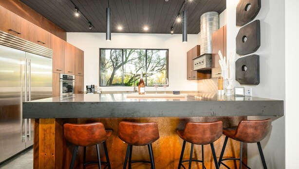 Une cuisine moderne dotée d’un grand îlot central, de tabourets de bar, d’appareils électroménagers en acier inoxydable et d’une grande fenêtre offrant une vue sur les arbres.