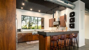 Une cuisine moderne dotée d’un grand îlot central, de tabourets de bar et offrant une vue sur les arbres grâce à de grandes fenêtres.
