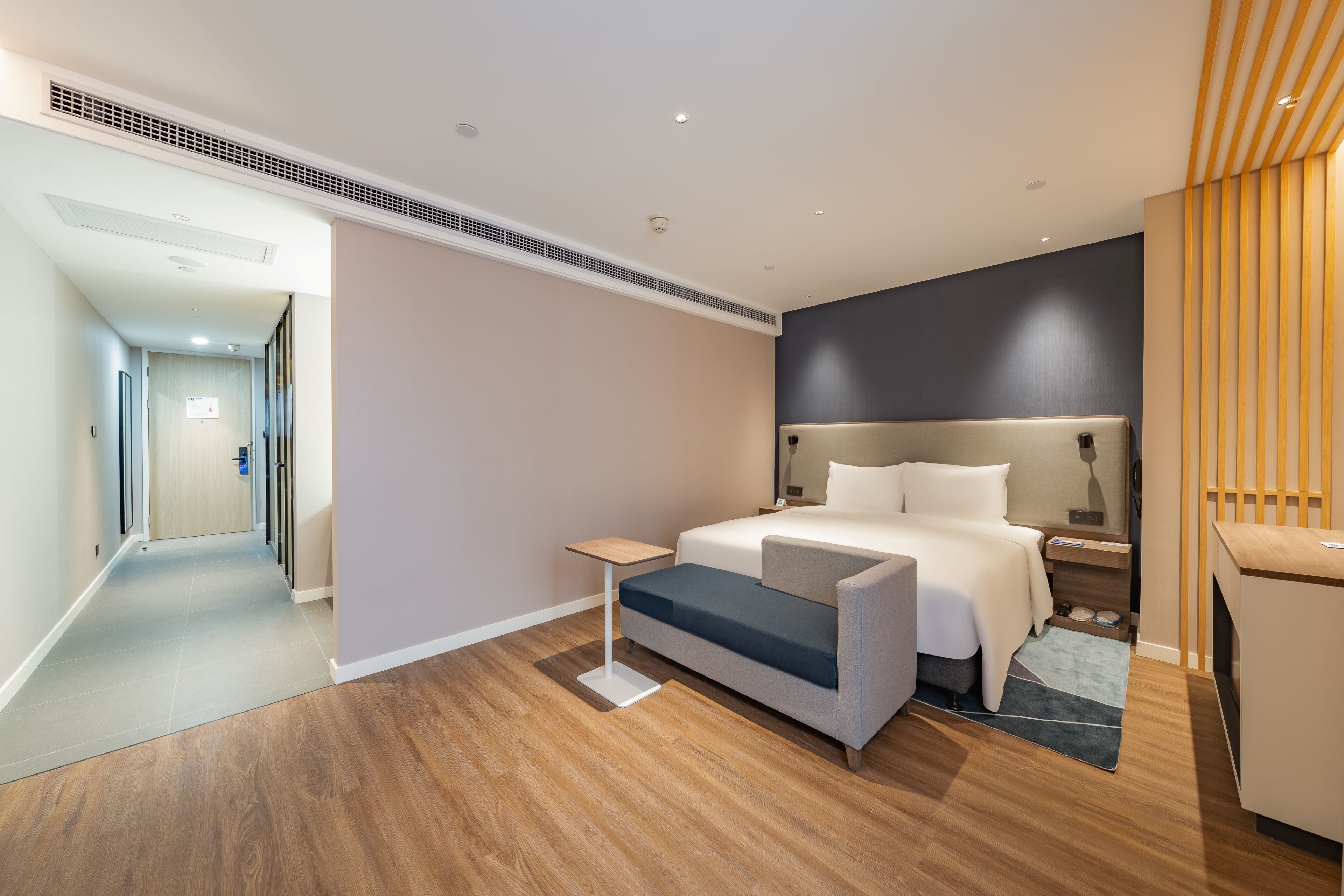 A modern hotel room with a bed, bedside table, and a desk. There is a hallway leading to another room.
