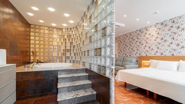 Una habitación de hotel moderna con piso de mármol, una cama grande, una zona de estar con sofá y un baño con pared de vidrio y bañera.