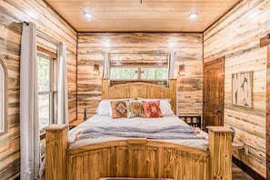 A wooden cabin bedroom with a bed, curtains, a window, and a door.
