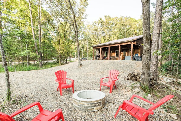 A cabin with a stone fire pit surrounded by red Adirondack chairs, set in a forested area with a gravel ground. - Calvert's Copper Cabin, Cozy cabin! Hot tub, Free Firewood, Fireplace! (Broken Bow)