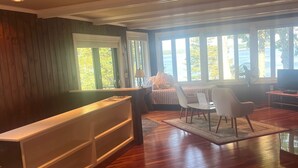 A spacious room with a large bed, a desk, and a chair. There are multiple windows providing a view of the outdoors.