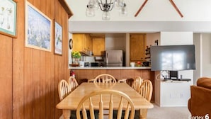 Un coin repas avec une table et des chaises en bois, une cuisine avec des meubles en bois, un réfrigérateur et un téléviseur à écran plat posé sur un meuble.