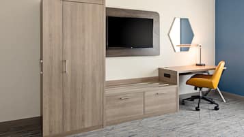 A modern room with a wooden wardrobe, a flat-screen TV mounted on the wall, a desk with a chair, and a mirror.