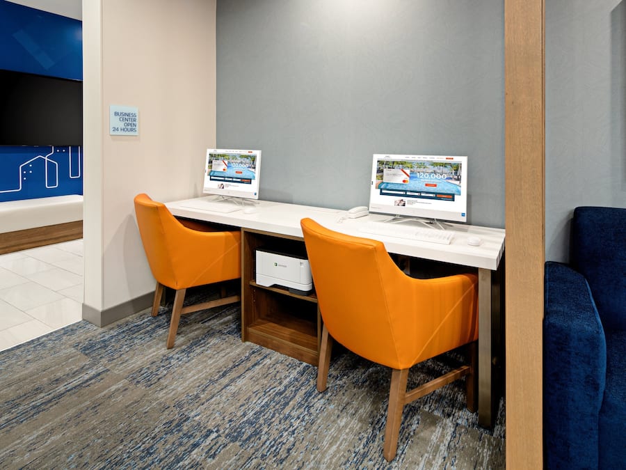 Un centre d’affaires équipé de deux postes informatiques, de chaises orange et d’un canapé bleu.