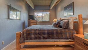 A bedroom with a wooden bed, a nightstand, a lamp, a TV, and a window with a view of houses and trees.