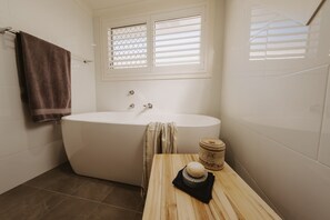 A bathroom with a freestanding bathtub, a window with blinds, a towel rack, and a small table with a soap dish.