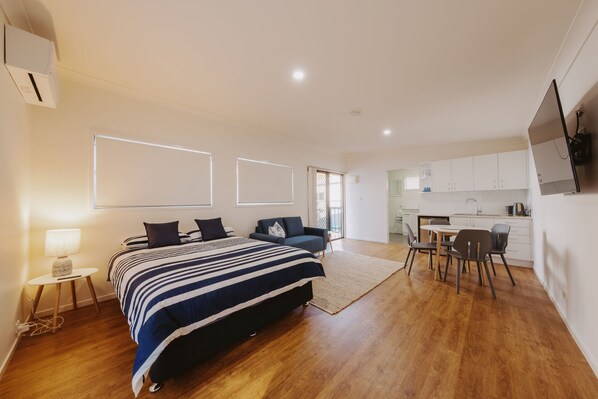 A modern bedroom with a large bed, bedside tables, a desk, and a kitchenette.