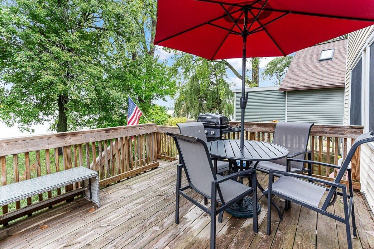 A wooden deck with a round table and chairs, a red umbrella, and an American flag.