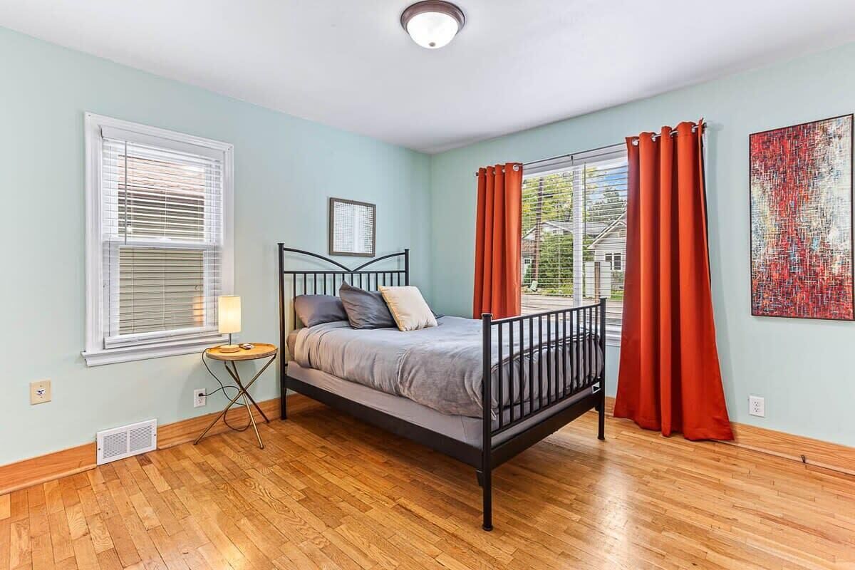 A bedroom with a bed, a window with blinds, a radiator, a small table with a lamp, and a painting on the wall.