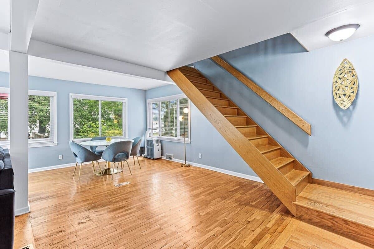 A spacious room with a wooden staircase, large windows, and a dining area with chairs.
