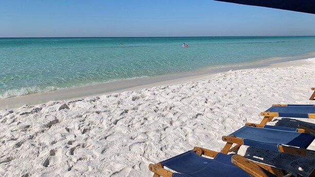 Een strand met wit zand en helder turquoise water. Er staan houten ligbedden onder een blauwe parasol.