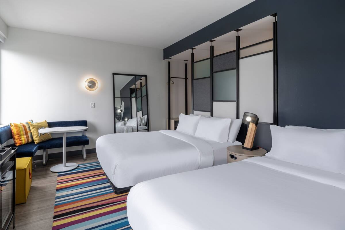 A hotel room with two beds, a small table, a sofa, a mirror, and a nightstand with a lamp.