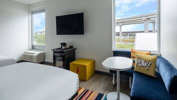 A hotel room with a bed, a desk, a TV, a small table, a sofa, and a window with a view of an overpass.