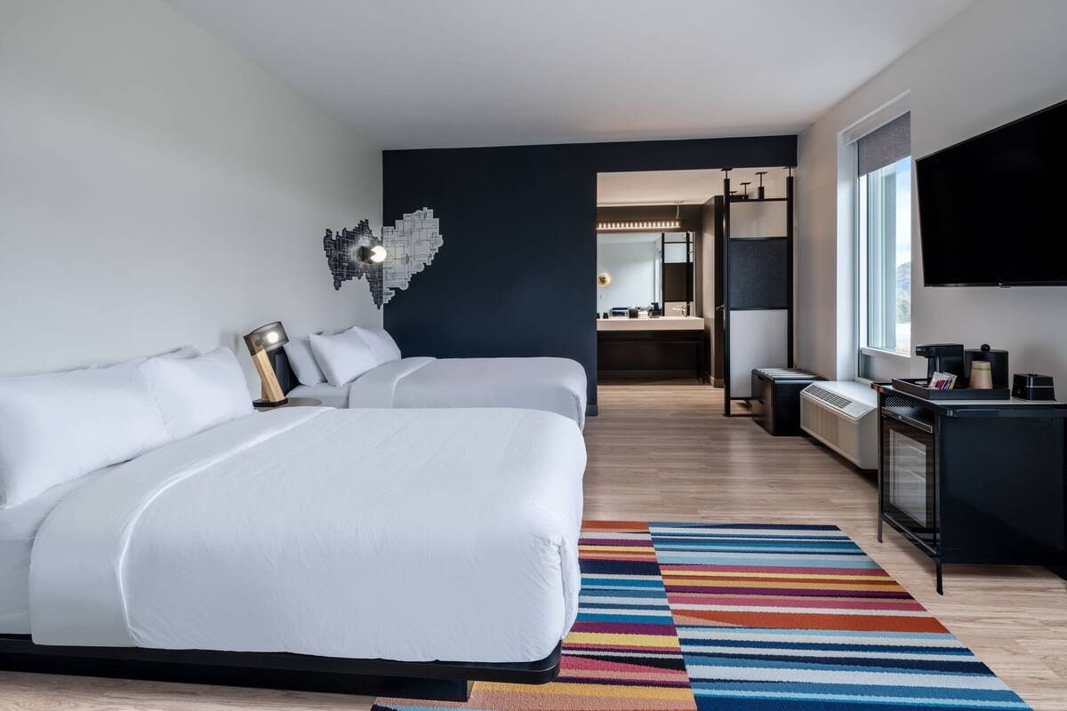 A modern hotel room with a large bed, a desk with a flat-screen TV, and a bathroom visible through an open door.