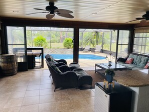 Une pièce spacieuse dotée d’une grande porte vitrée donnant sur un jardin avec une piscine, du mobilier d’extérieur et un ventilateur de plafond.