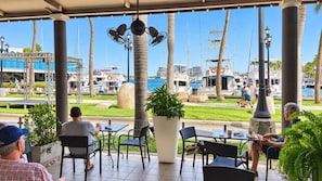 Ein überdachter Außenbereich mit Tischen und Stühlen, mit Blick auf einen Yachthafen und Boote.