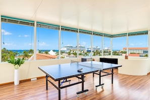 Ein geräumiges Zimmer mit einem Pingpong-Tisch, großen Fenstern mit Blick auf das Meer und Kreuzfahrtschiffe sowie einer Zimmerpflanze.