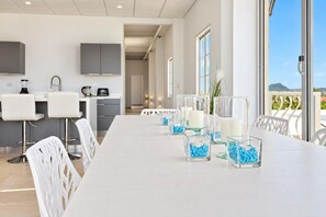 A modern dining area with a long table, white chairs, and clear glass candle holders with blue pebbles.