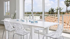 Un vaste balcon doté d’une table et de chaises blanches, offrant une vue sur un paysage côtier avec des palmiers et des bâtiments.