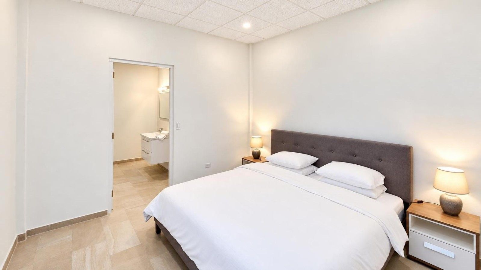 A hotel room with a bed, bedside tables, a nightstand, a lamp, and a bathroom visible through an open door.
