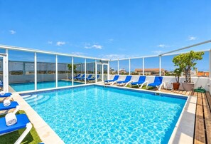 A rooftop pool with clear blue water, surrounded by white-framed glass panels, and blue lounge chairs.