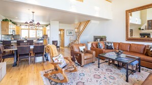 A spacious living area with a large brown leather sofa, a glass coffee table, and a wooden rocking chair. A staircase leads to an upper level, and there is a kitchen area visible in the background.