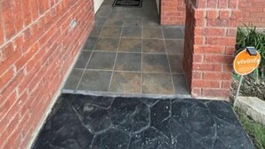 Um edifício de tijolos com uma porta branca, entrada com azulejos e uma pequena placa à direita.