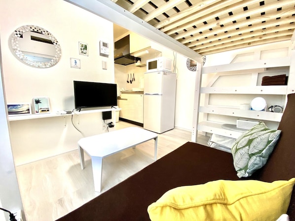 A compact living space with a flat-screen TV, a bench, a small table, and a kitchenette in the background.