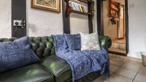 A green leather sofa with a blue blanket and pillows, a wooden bookshelf with books, and a framed picture on the wall. - Runnymede (Tewkesbury)