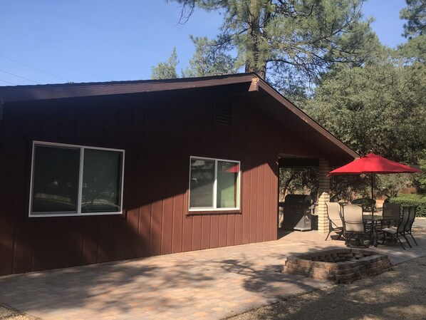 A wooden cabin with two windows, a patio with a red umbrella, and a fire pit.