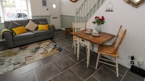 A living room with a grey sofa, yellow cushions, a wooden dining table with chairs, a vase of red flowers, and a staircase with white railings. - Y Bwthyn (Conwy)